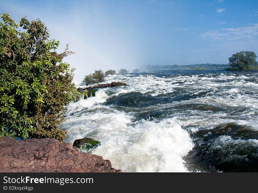 The raging torrent of the mighty Zambezi river in full flood at the very edge of the Victoria Falls. The spray cloud creates a subtle rainbow. The raging torrent of the mighty Zambezi river in full flood at the very edge of the Victoria Falls. The spray cloud creates a subtle rainbow.