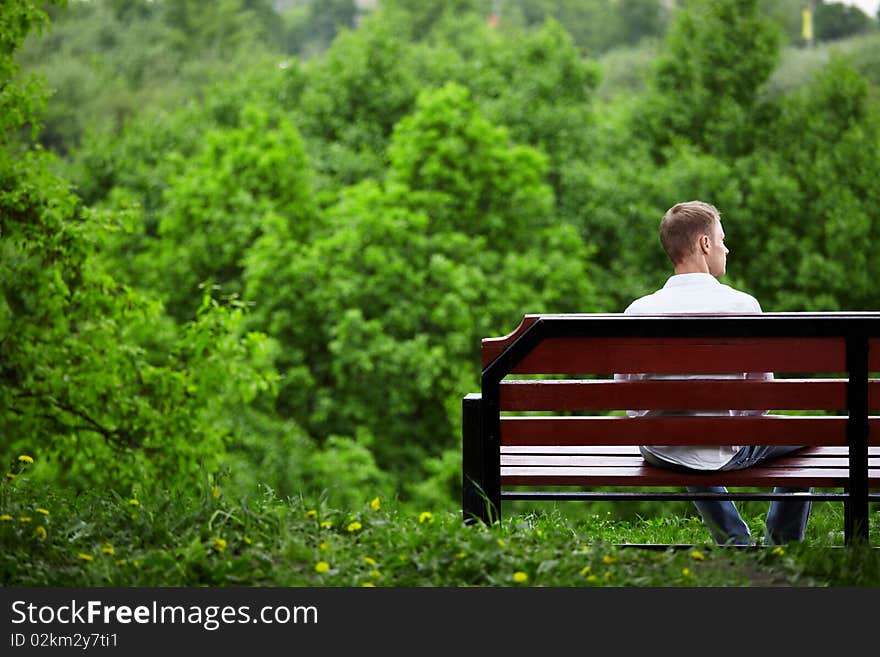 The young man sits one on a bench against green trees. The young man sits one on a bench against green trees