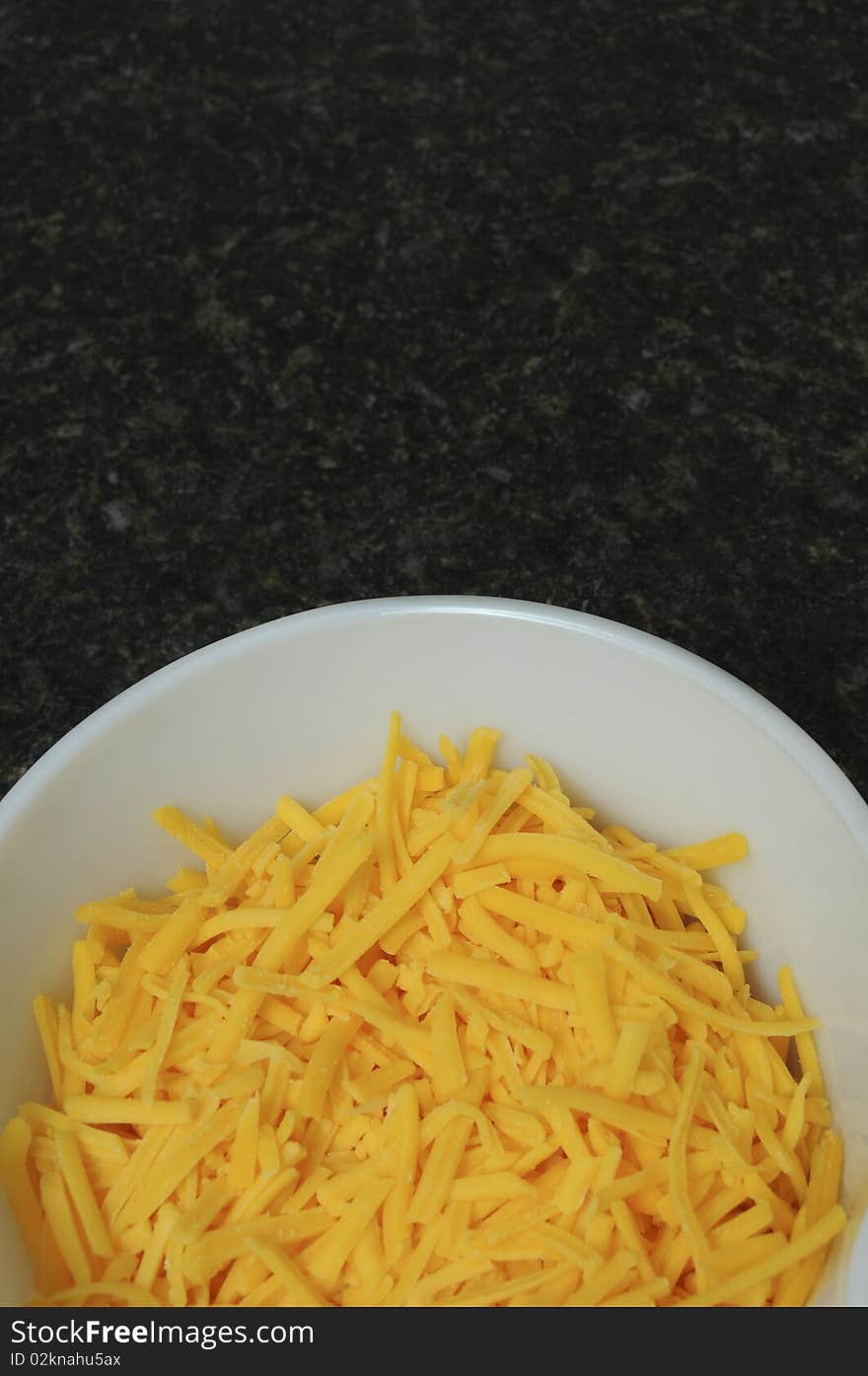 An aerial view of a white bowl containing yellow cheddar cheese on a black counter top. An aerial view of a white bowl containing yellow cheddar cheese on a black counter top