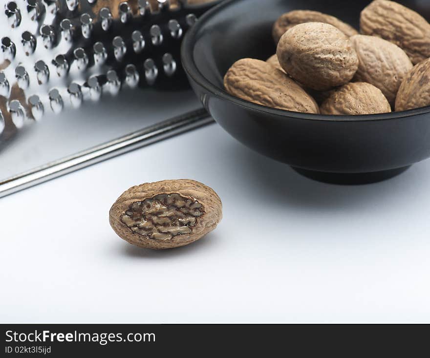 A Part Grated Nutmeg, In Front Of A Bowl Of Nutmegs and A Grater, On A White Background