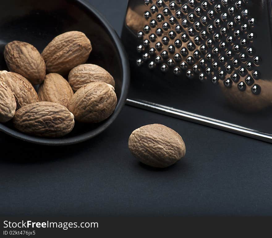 A Nutmeg On A Black Background, In Front Of A Black Dish Of Nutmegs and A Chrome Grater