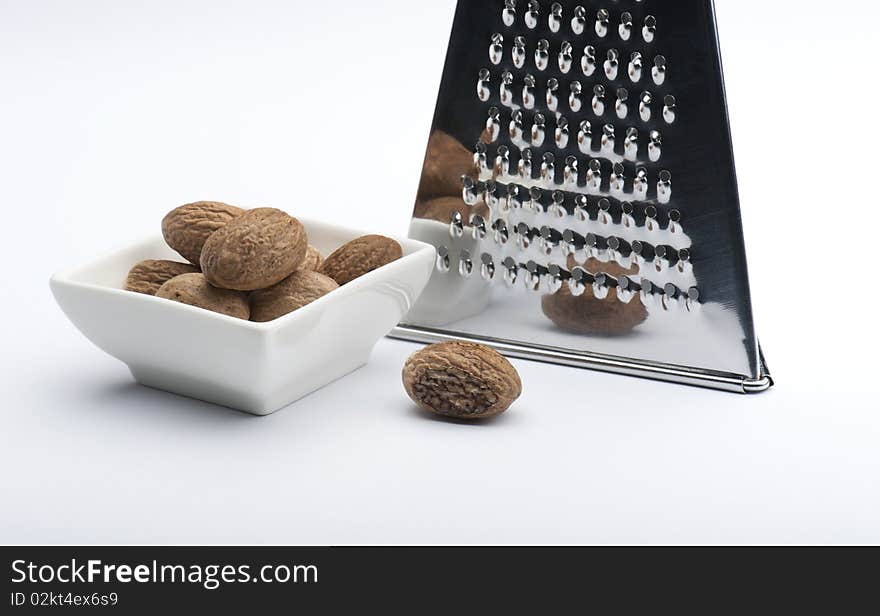 A Partly Grated Nutmeg In Front Of A Chrome Grater and Next To A White Dish Full Of Nutmegs, On A White Background