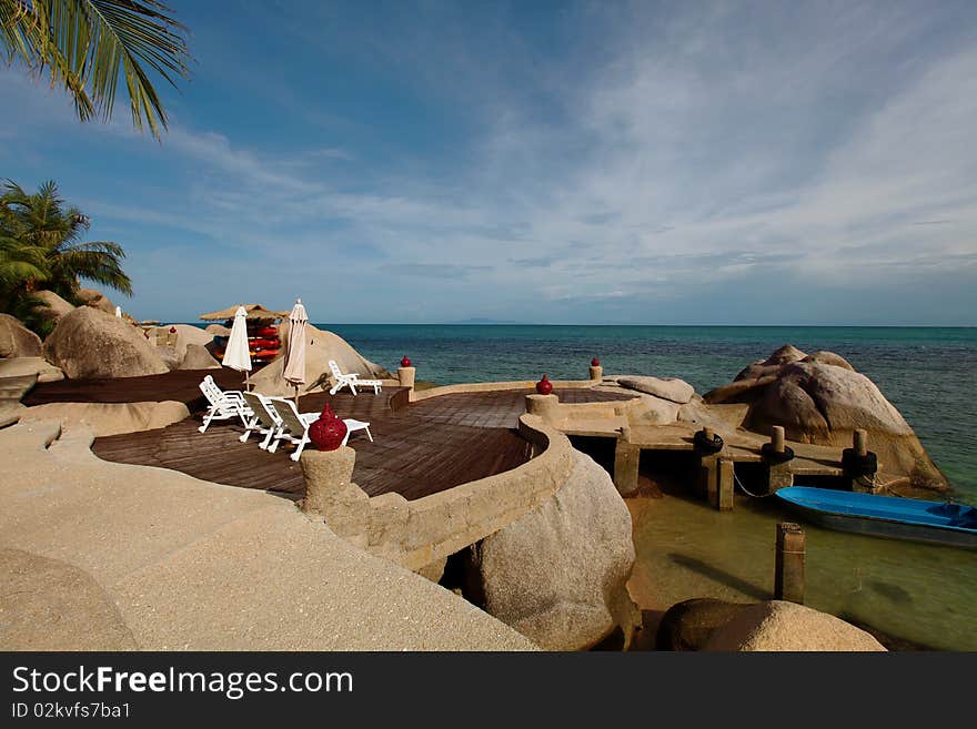 A little private pier of a resort in Koh Tao island, Thailand, near a sundeck on the rocks, with lounge chairs and a hut with canoes for guests. A little private pier of a resort in Koh Tao island, Thailand, near a sundeck on the rocks, with lounge chairs and a hut with canoes for guests.