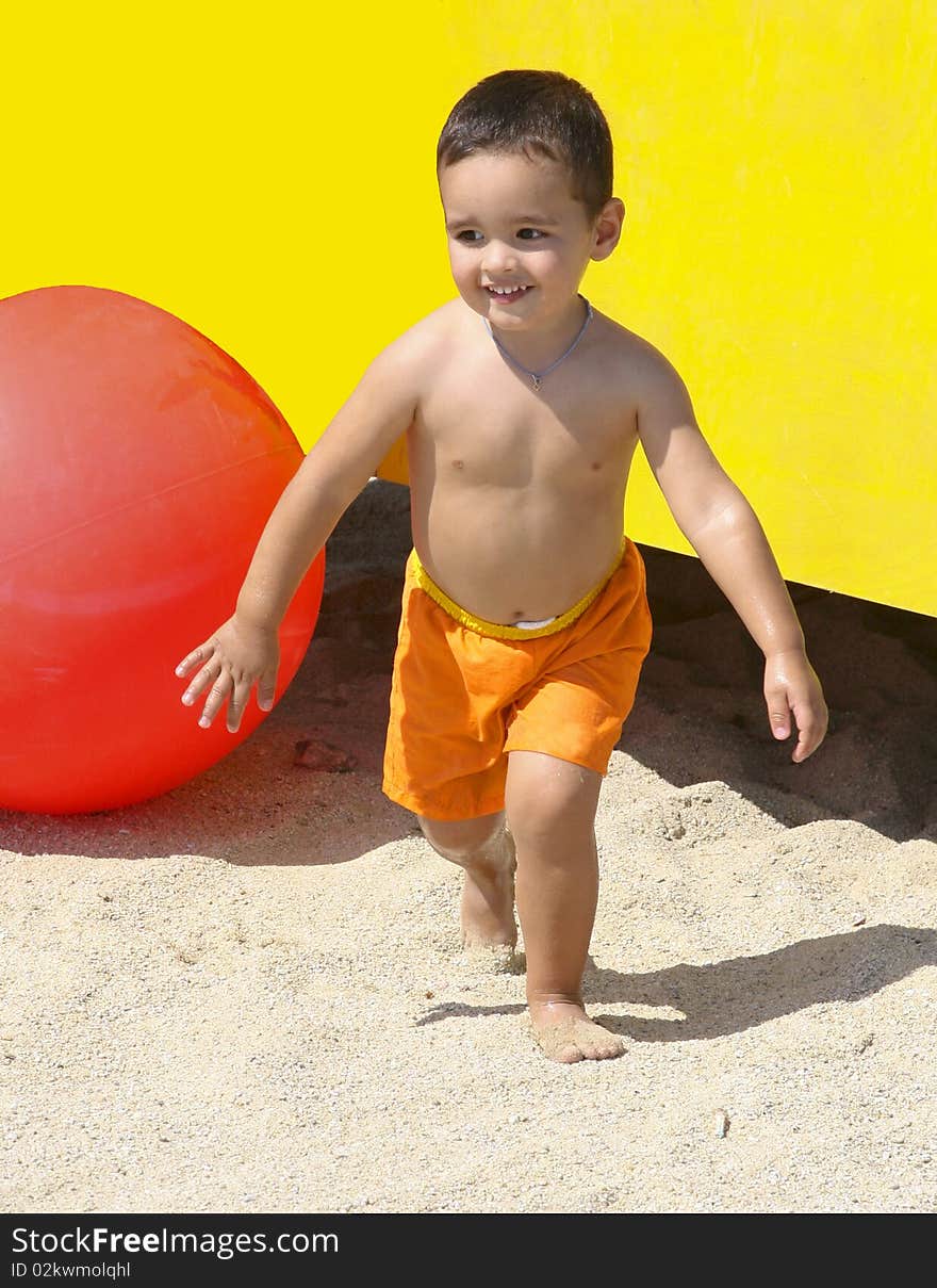 A happy little boy in an an orange swim suit, running surrounded by a huge red ball , yellow background and sand. A happy little boy in an an orange swim suit, running surrounded by a huge red ball , yellow background and sand.