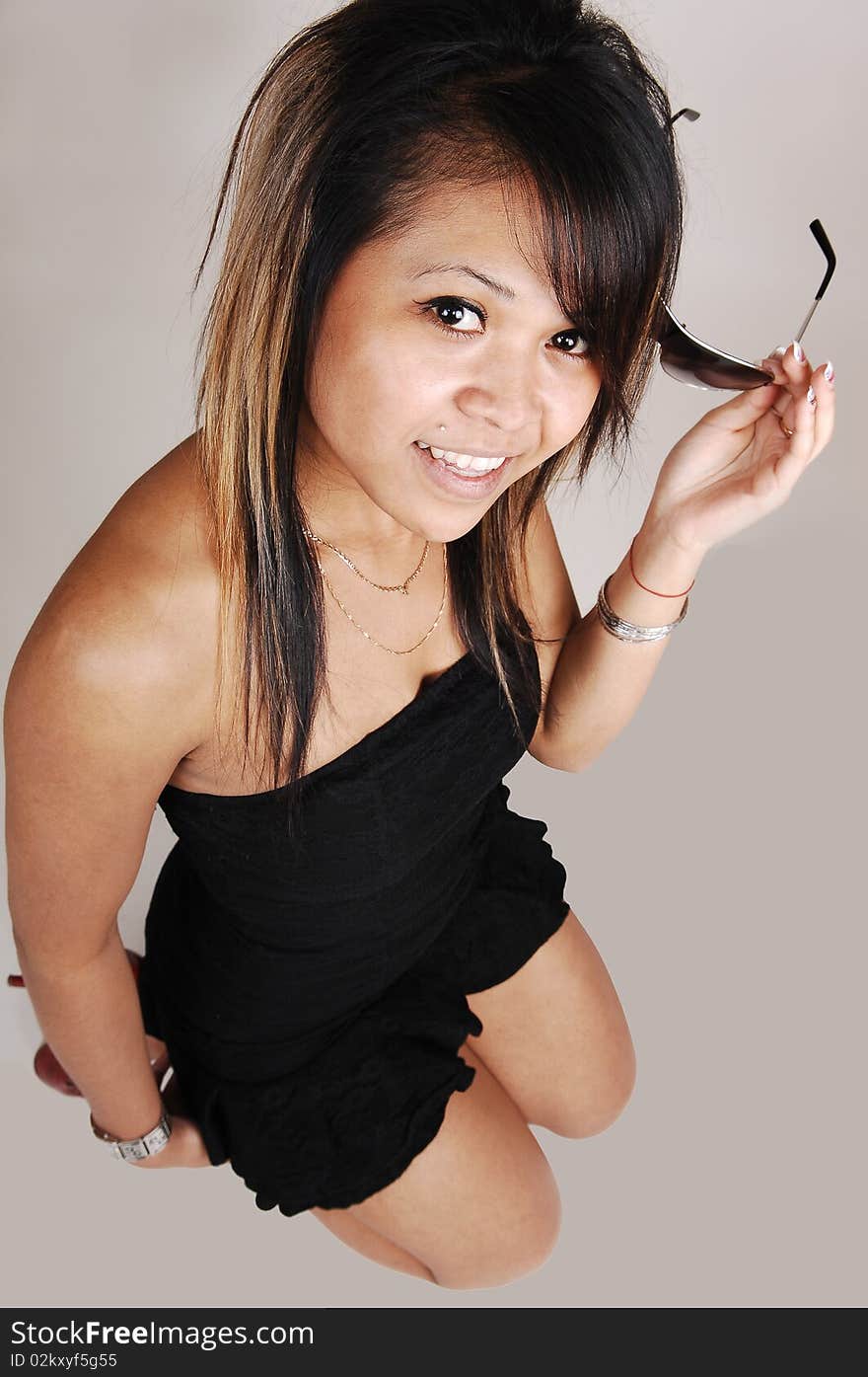 A young pretty Asian woman, in a short black dress kneeling on the floor in the studio and smiling in the camera, on light gray background. A young pretty Asian woman, in a short black dress kneeling on the floor in the studio and smiling in the camera, on light gray background.