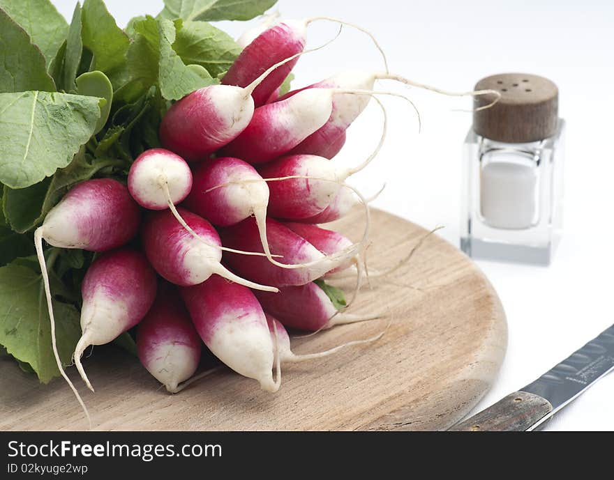 A Bunch Of Fresh French Breakfast Radish On A Wooden Chopping Board, With A Salt Pot and knife, On A White Background. A Bunch Of Fresh French Breakfast Radish On A Wooden Chopping Board, With A Salt Pot and knife, On A White Background