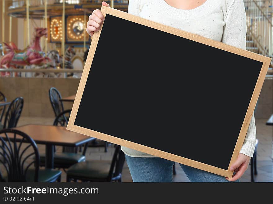 Female holding blank chalkboard in front of carousel in mall food court. Female holding blank chalkboard in front of carousel in mall food court.