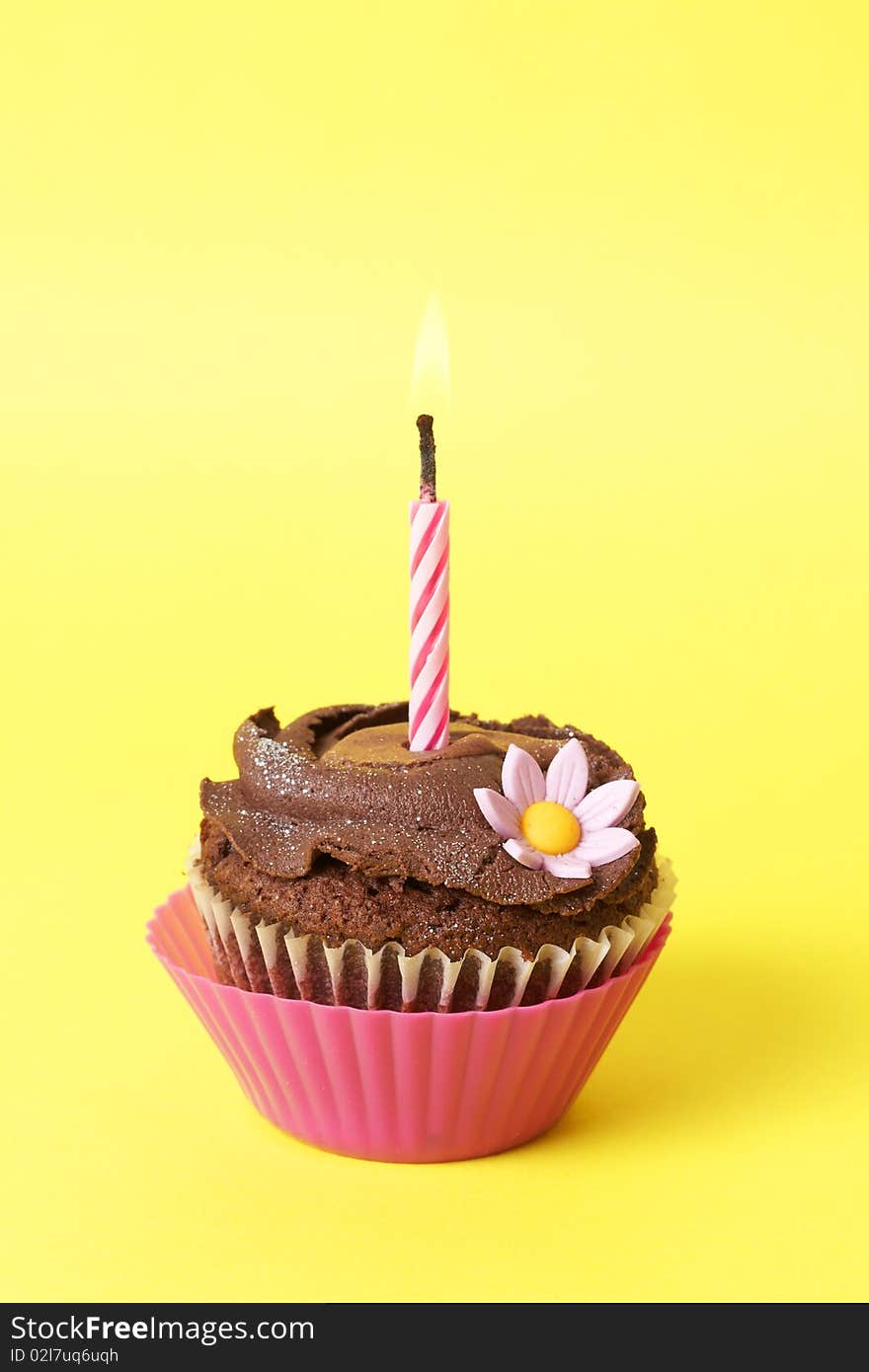 Miniature chocolate cupcake with icing, decorative flower and birthday candle on yellow background. Miniature chocolate cupcake with icing, decorative flower and birthday candle on yellow background