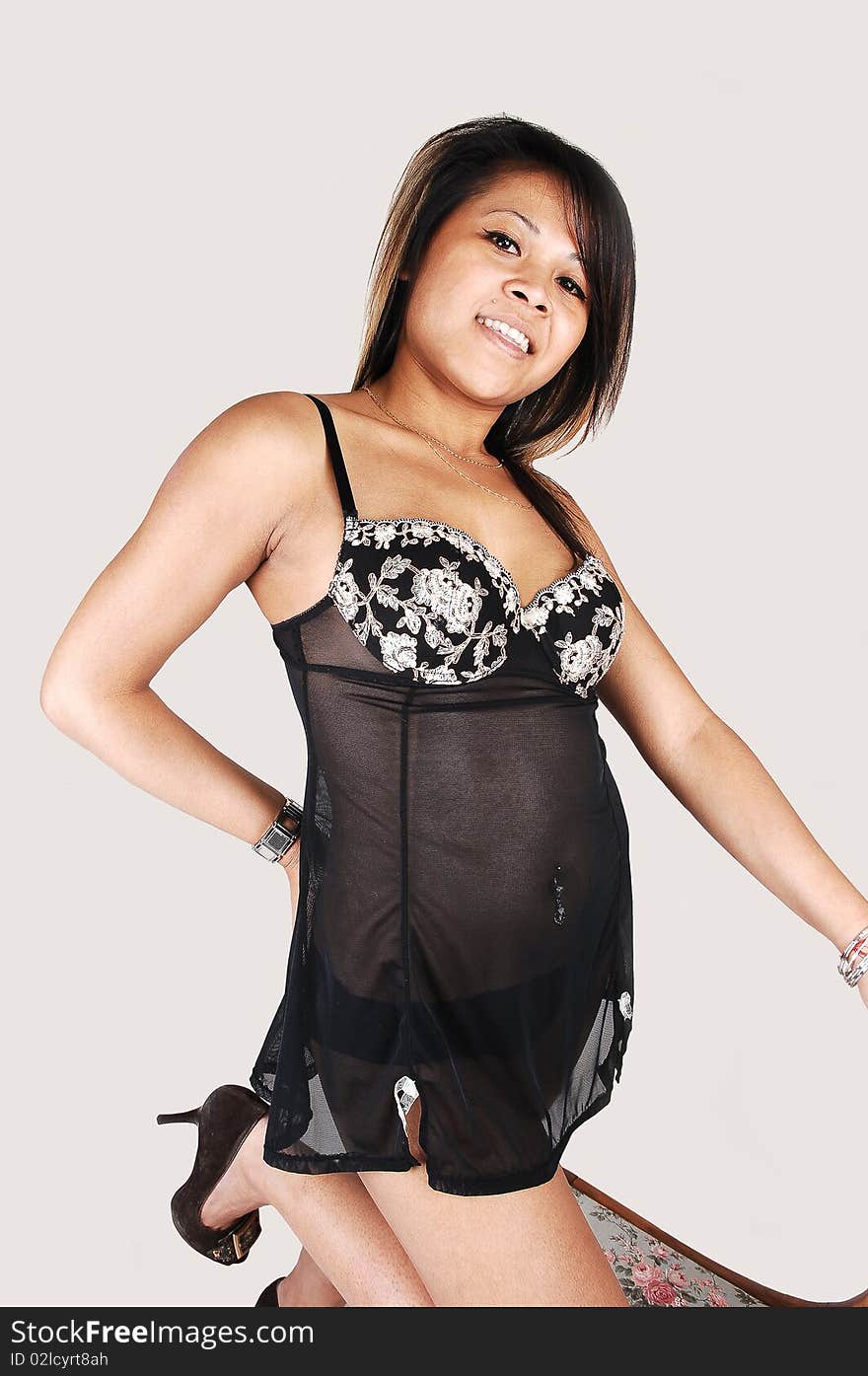 A young Asian woman in black lingerie kneeling with one knee on an chair smiling into the camera, over light gray background. A young Asian woman in black lingerie kneeling with one knee on an chair smiling into the camera, over light gray background.