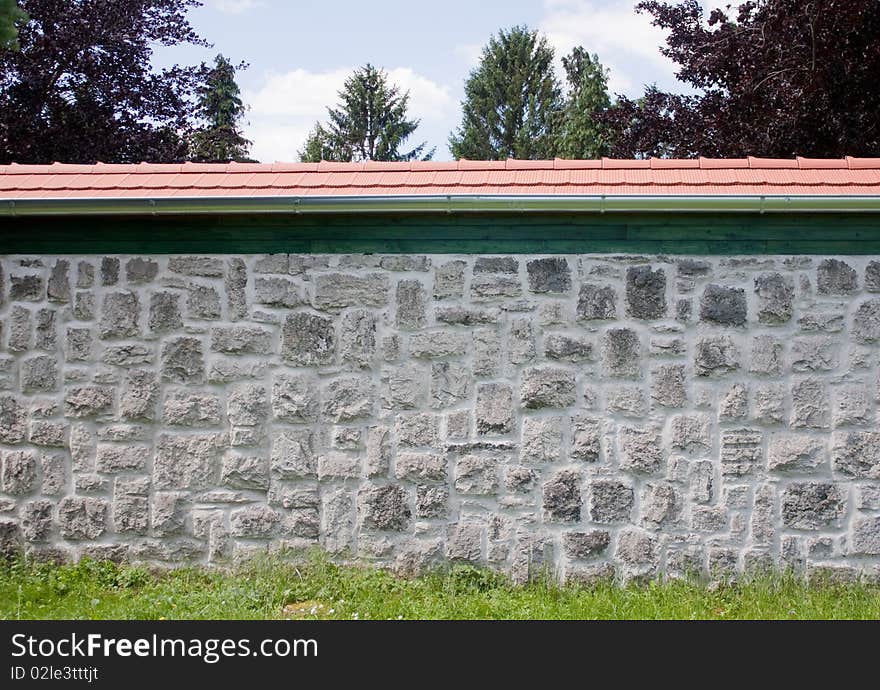 A stone wall with some grass in the foreground and with red roof, with some trees visible beyond the wall. A stone wall with some grass in the foreground and with red roof, with some trees visible beyond the wall