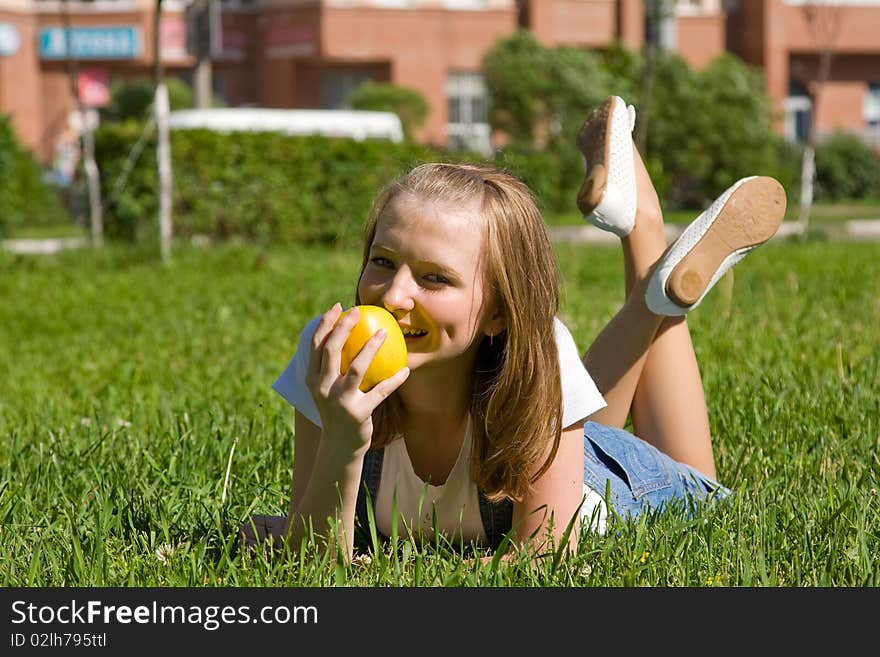 Teenage girl with a yellow apple. Beautiful young woman lying on a green grass and eating apple outside on field.