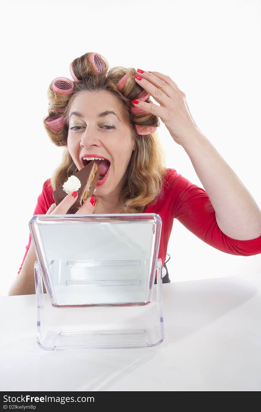 Middle-aged woman with hair rollers eating cake looking at mirror