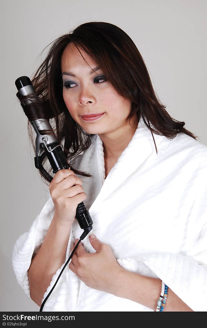 A pretty Chinese woman, is curling her hair, dressed in a white bath robe for light gray background. A pretty Chinese woman, is curling her hair, dressed in a white bath robe for light gray background.