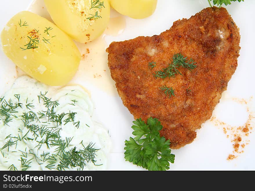 Pork chop (covered in batter and breadcrumbs), potatoes and cucumber salad decorated with dill and parsley on a plate on white background. Pork chop (covered in batter and breadcrumbs), potatoes and cucumber salad decorated with dill and parsley on a plate on white background