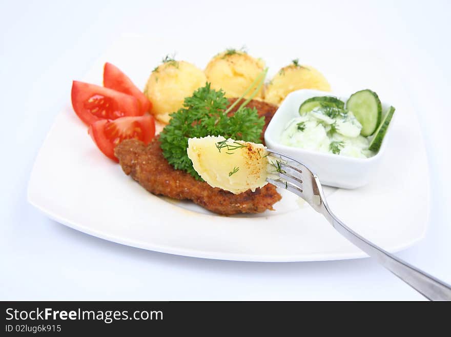 Pork chop (covered in batter and breadcrumbs), mashed potatoes-some of it on a fork, cucumber salad in a bowl and tomato pieces decorated with dill and parsley on a plate with a on white background. Pork chop (covered in batter and breadcrumbs), mashed potatoes-some of it on a fork, cucumber salad in a bowl and tomato pieces decorated with dill and parsley on a plate with a on white background