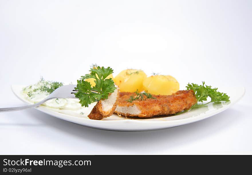 Pork chop (covered in batter and breadcrumbs)- piece of it on a fork- potatoes and cucumber salad decorated with dill and parsley on a plate on white background. Pork chop (covered in batter and breadcrumbs)- piece of it on a fork- potatoes and cucumber salad decorated with dill and parsley on a plate on white background