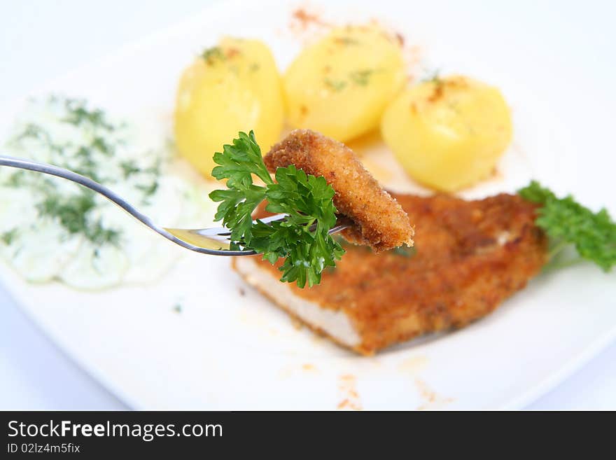 Pork chop (covered in batter and breadcrumbs)- piece of it on a fork- potatoes and cucumber salad decorated with dill and parsley on a plate on white background. Pork chop (covered in batter and breadcrumbs)- piece of it on a fork- potatoes and cucumber salad decorated with dill and parsley on a plate on white background