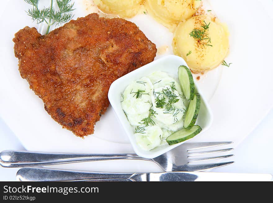 Pork chop (covered in batter and breadcrumbs), mashed potatoes and cucumber salad in a bowl decorated with dill on a plate with a fork and a knife on white background. Pork chop (covered in batter and breadcrumbs), mashed potatoes and cucumber salad in a bowl decorated with dill on a plate with a fork and a knife on white background