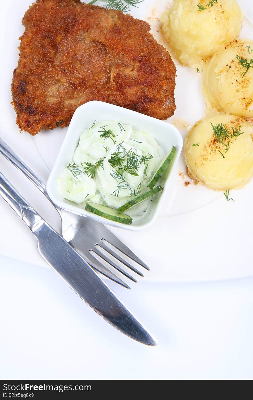 Pork chop (covered in batter and breadcrumbs), mashed potatoes and cucumber salad in a bowl decorated with dill on a plate with a fork and a knife on white background. Pork chop (covered in batter and breadcrumbs), mashed potatoes and cucumber salad in a bowl decorated with dill on a plate with a fork and a knife on white background