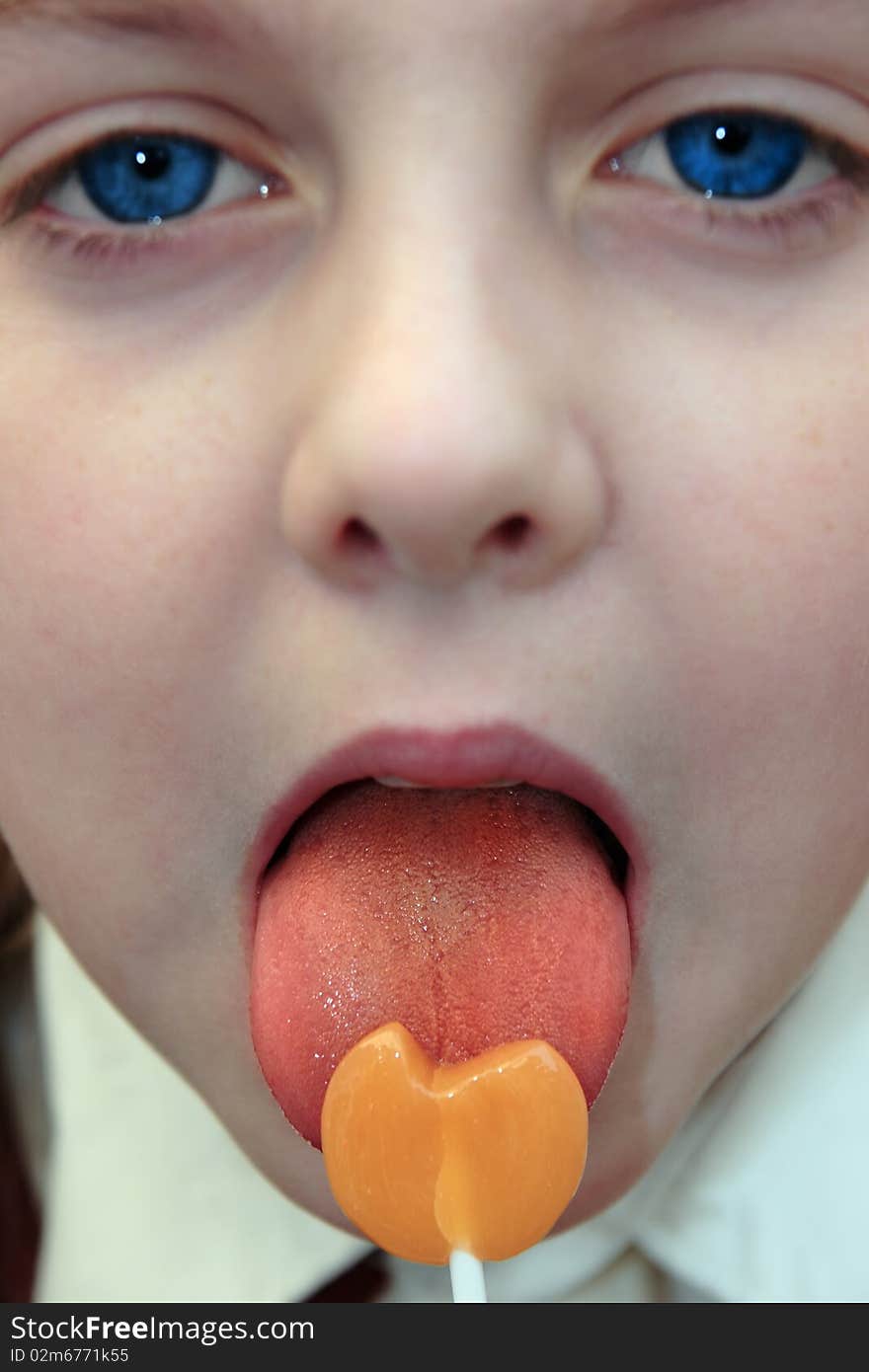 Young blue eyed girl licking an orange lollipop into a heart shape