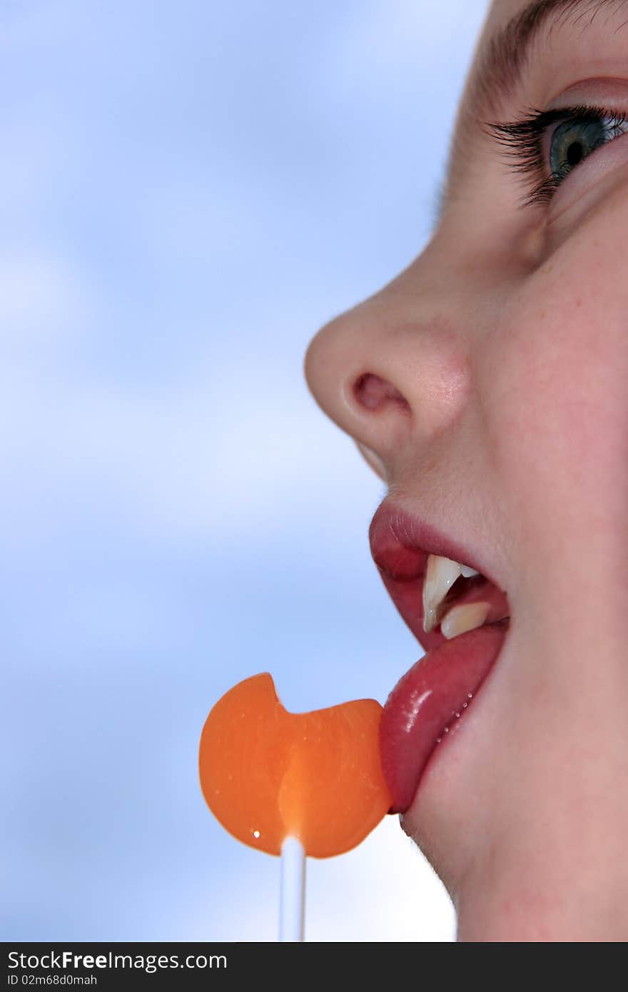 Young blue eyed girl licking an orange lollipop into a heart shape against a blue sky. Young blue eyed girl licking an orange lollipop into a heart shape against a blue sky