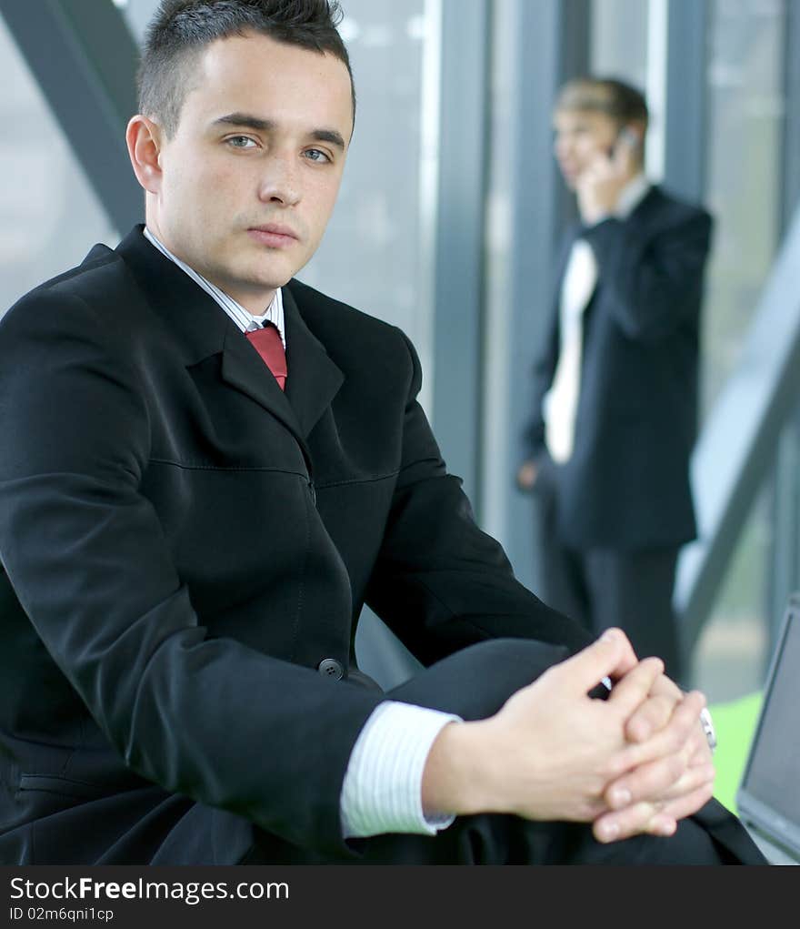 Portrait of a young business man in formal clothes sitting in an office. Another business man is talking to the mobile phone on the background. Portrait of a young business man in formal clothes sitting in an office. Another business man is talking to the mobile phone on the background.