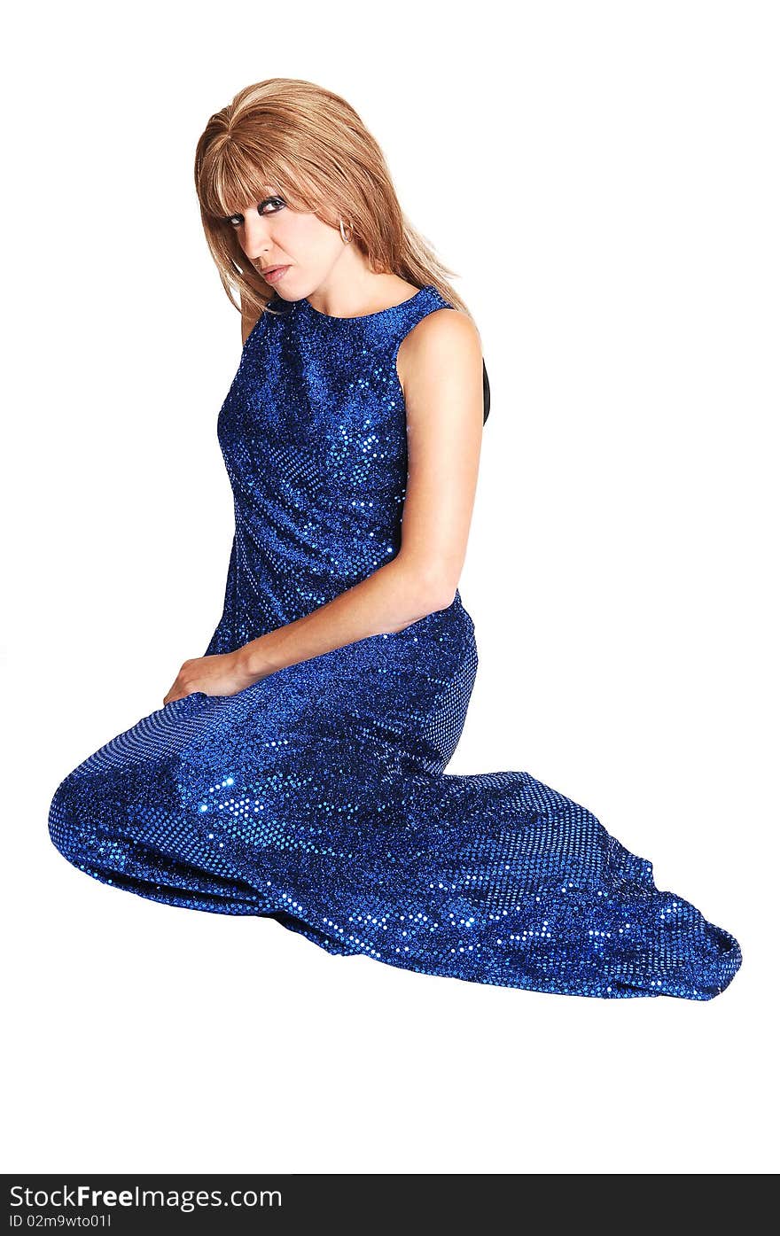 A brunette young pretty woman in a long navy blue dress sitting on the floor in the studio looking shy into the camera on white background. A brunette young pretty woman in a long navy blue dress sitting on the floor in the studio looking shy into the camera on white background.