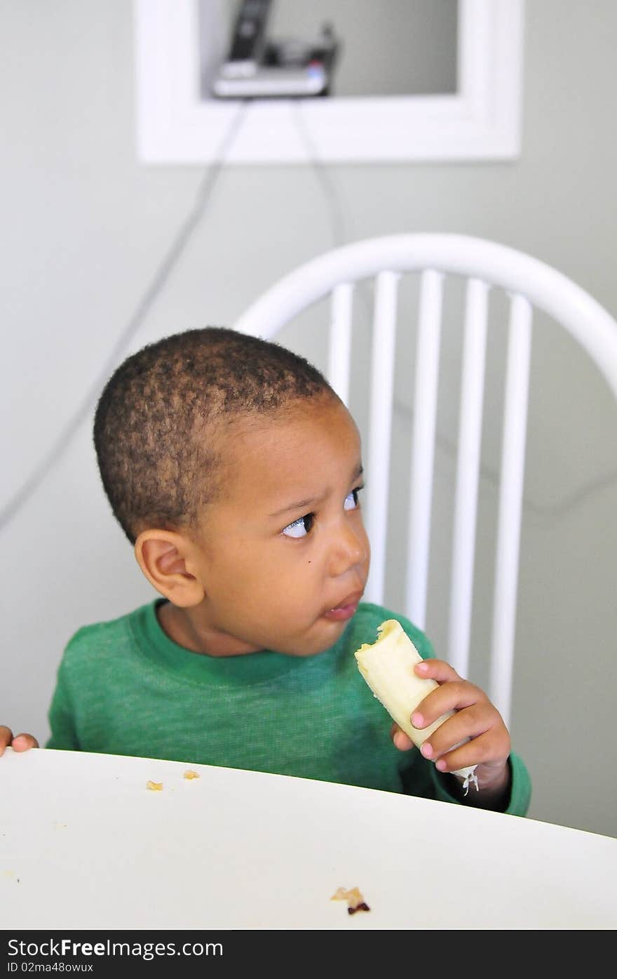 Young child sitting at table with banana in hand eating while looking away. Young child sitting at table with banana in hand eating while looking away