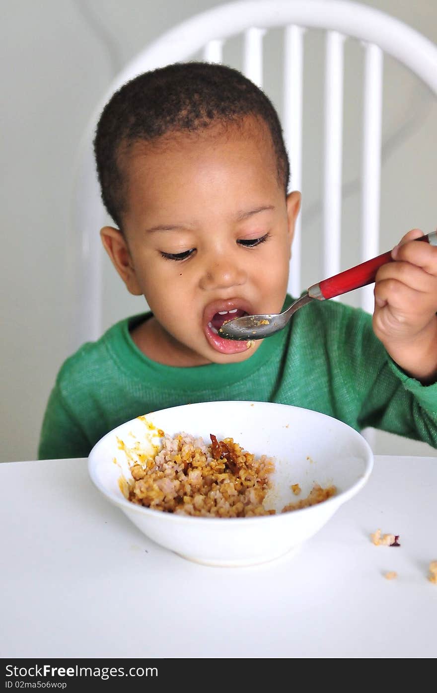 Young boy eating at table from white dish with red spoon. Young boy eating at table from white dish with red spoon