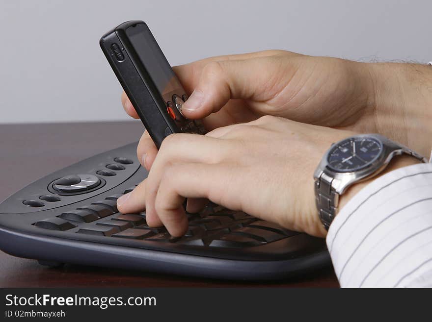 A man working on a computer, holding a cellular phone in one hand. A man working on a computer, holding a cellular phone in one hand