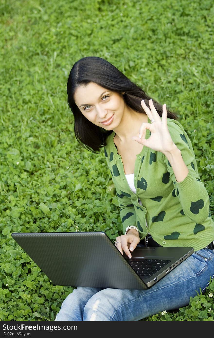 Young Caucasian woman with a laptop in the park on a green grass / meadow. Sign OK