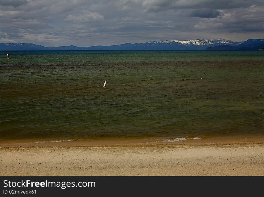 A sandy beach on Lake Tahoe Beach, Nevada.  The mountains are in the background and is a cloudy day.  The water has hues of green, brown and yellow.