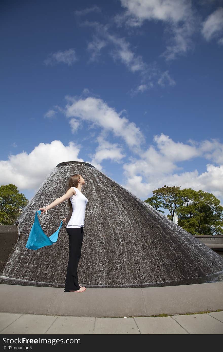 A woman holding her blue sarong while standing near a beautiful water fountain letting the breeze blow her sarong. A woman holding her blue sarong while standing near a beautiful water fountain letting the breeze blow her sarong.