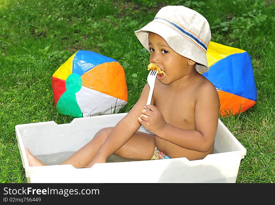 Young child eating while sitting in water with beech balls lying around him on grass. Young child eating while sitting in water with beech balls lying around him on grass