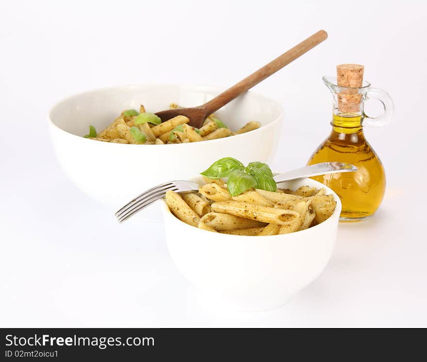 Penne with pesto decorated with basil and a fork in a small bowl and and a big bowl with wooden ladle , and a bottle of olive oil