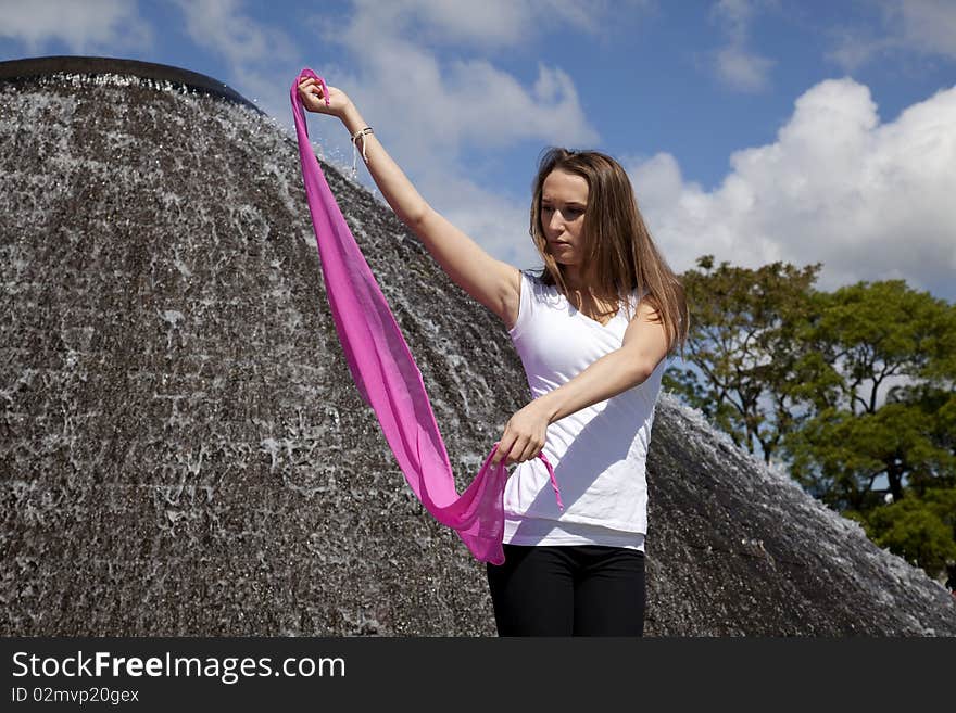 A woman holding and looking at her pink sarong with a longing expression her face, while she is standing in the park with a beautiful water fountain behind her. A woman holding and looking at her pink sarong with a longing expression her face, while she is standing in the park with a beautiful water fountain behind her.