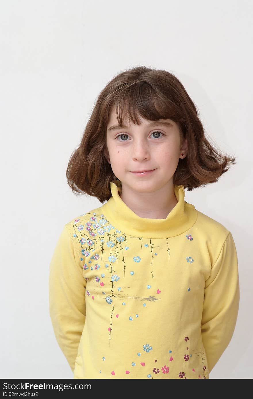 Serious cute little girl stands against the white wall with hands behind her back. Serious cute little girl stands against the white wall with hands behind her back