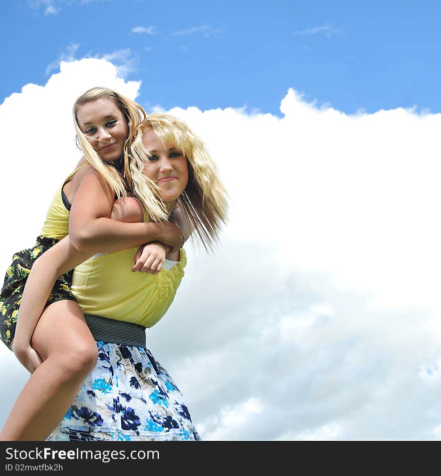 Two beautiful and attractive blond teenage girls giving a piggyback ride in the sunshine with clouds and sky in the background. Two beautiful and attractive blond teenage girls giving a piggyback ride in the sunshine with clouds and sky in the background.