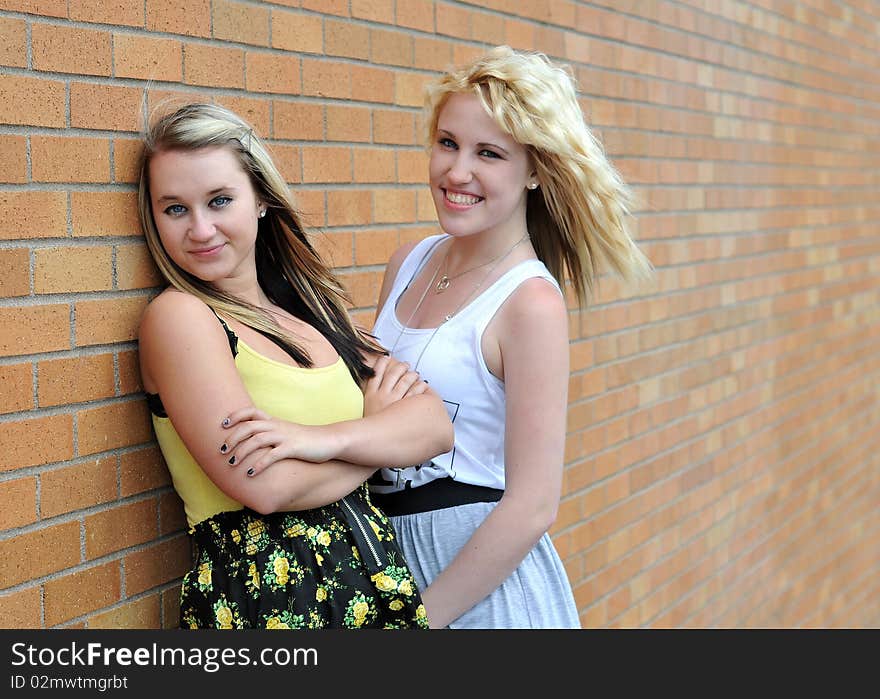 Happy young blond teenage girl friends standing in front of a brick wall smiling with each other. Happy young blond teenage girl friends standing in front of a brick wall smiling with each other.