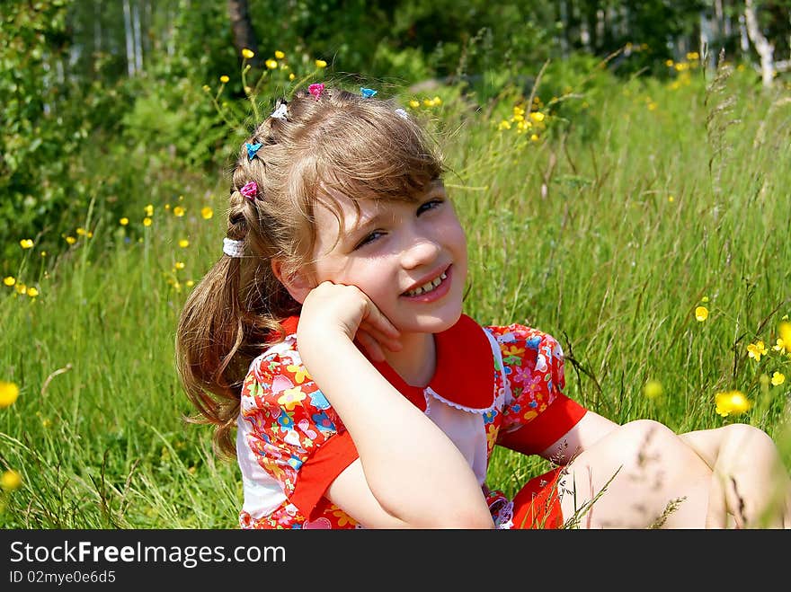 In the summer in park on a green grass with yellow flowers the smiling girl in a red dress sits. In the summer in park on a green grass with yellow flowers the smiling girl in a red dress sits