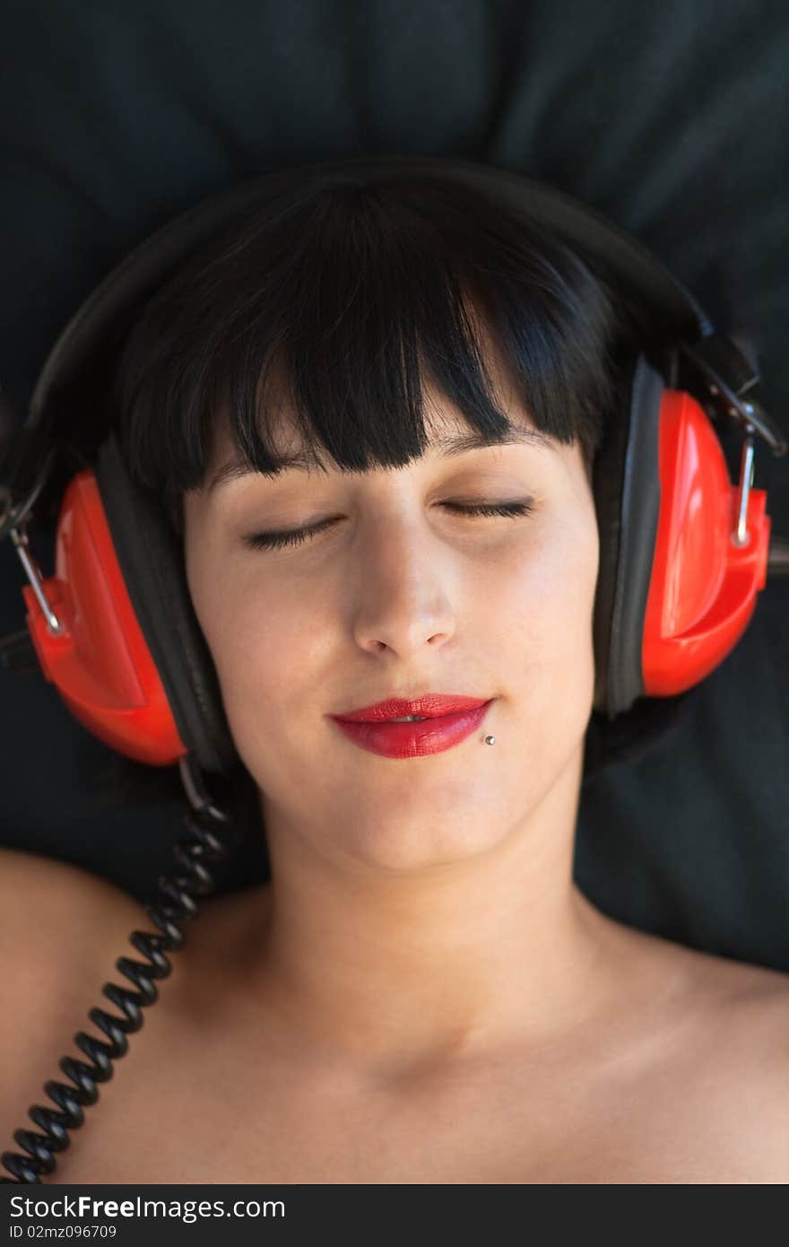 Beautiful women rests her head against a black pillow while enjoying music through her red head phones, relaxing. Beautiful women rests her head against a black pillow while enjoying music through her red head phones, relaxing.