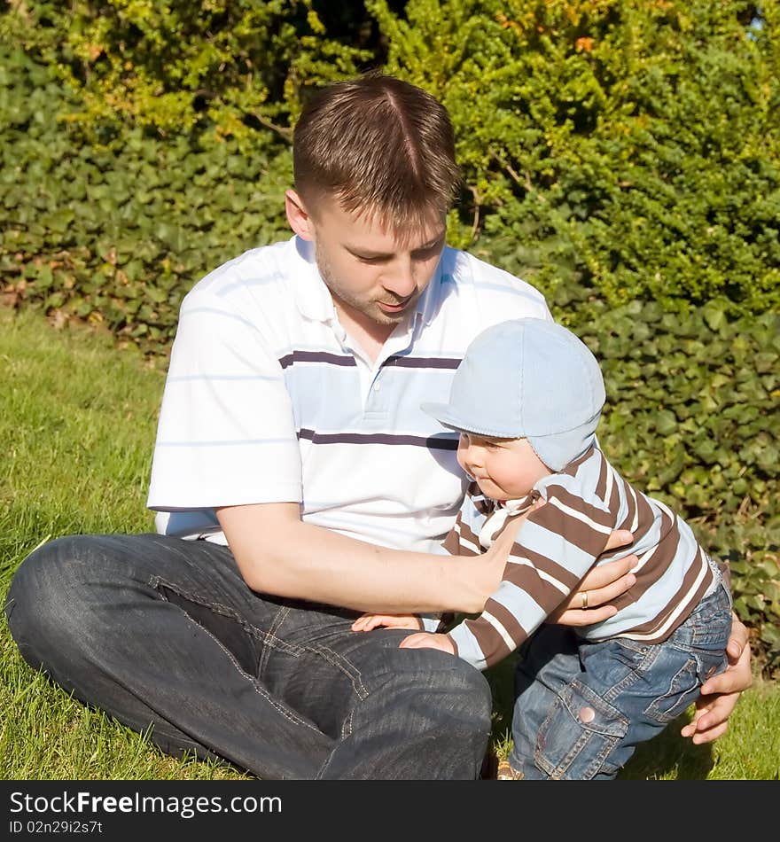 Father sitting with his son in park on the grass. Child puts first steps outside. Father secures his child. Father sitting with his son in park on the grass. Child puts first steps outside. Father secures his child.