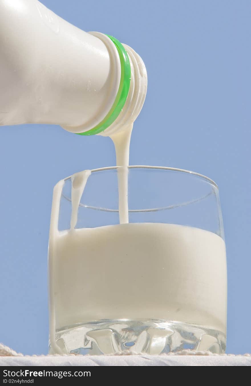 Glass from transparent glass with milk which is poured from a bottle on a blue background