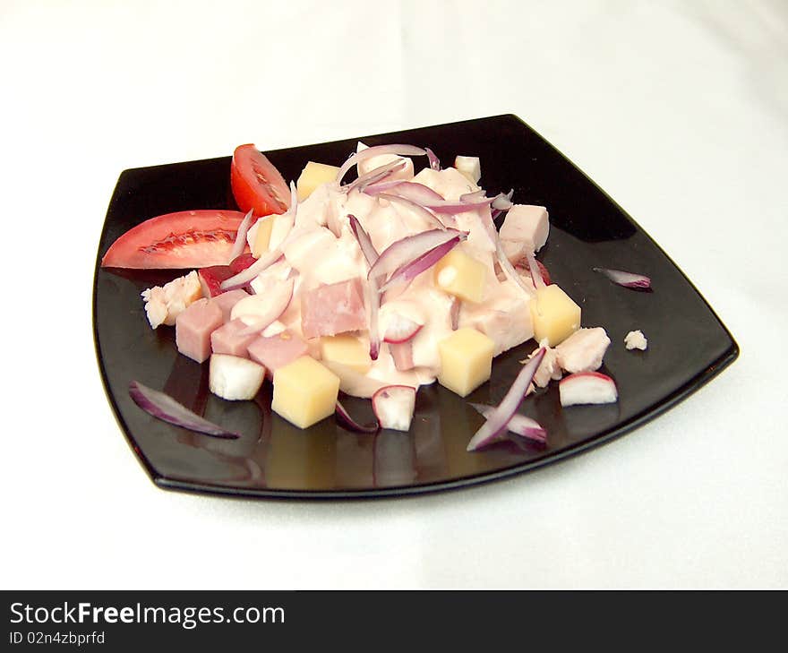 Salad with ham, cheese, redisos, chicken under cognac sauce, decorated with slices of tomato on a black square plate. Salad with ham, cheese, redisos, chicken under cognac sauce, decorated with slices of tomato on a black square plate