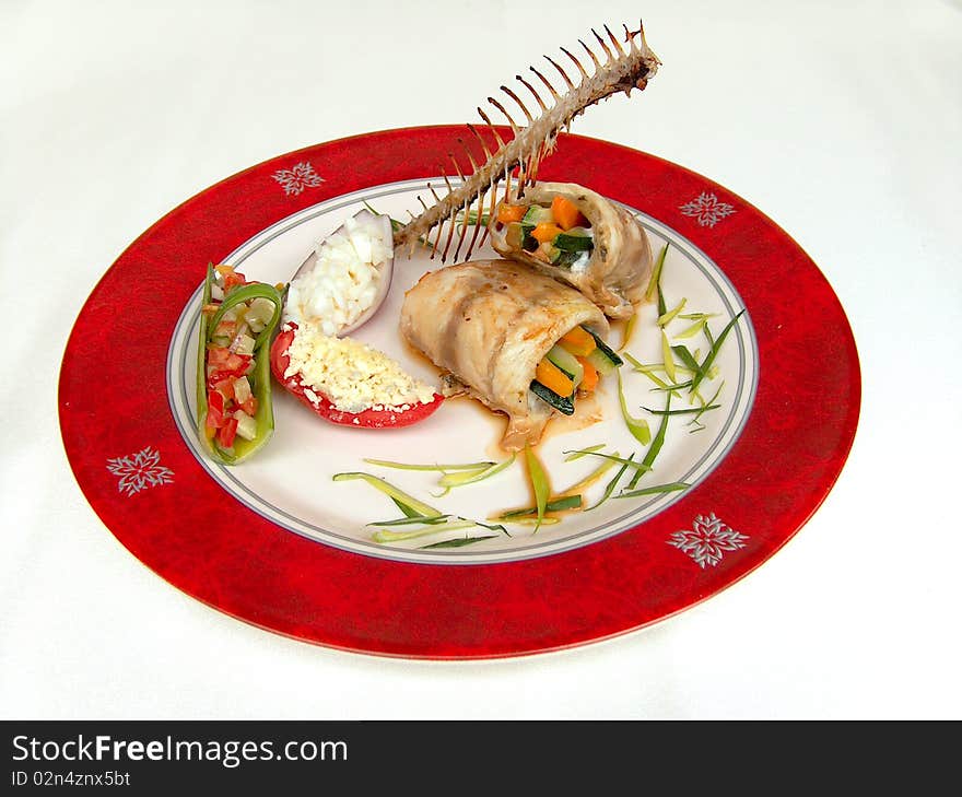 Fillet of seabass simmered with vegetables on the red plate with red border. This fish dish is decorate a fishbone. Fillet of seabass simmered with vegetables on the red plate with red border. This fish dish is decorate a fishbone.