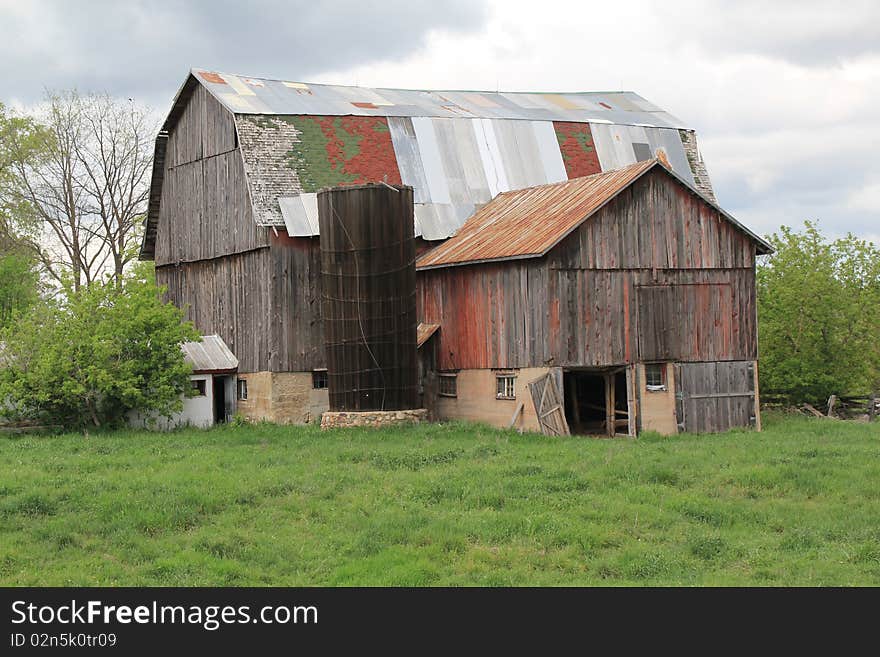 Old wood and metal barn in field with trees and sky background. Barn has many colors on roof. Old wood and metal barn in field with trees and sky background. Barn has many colors on roof.