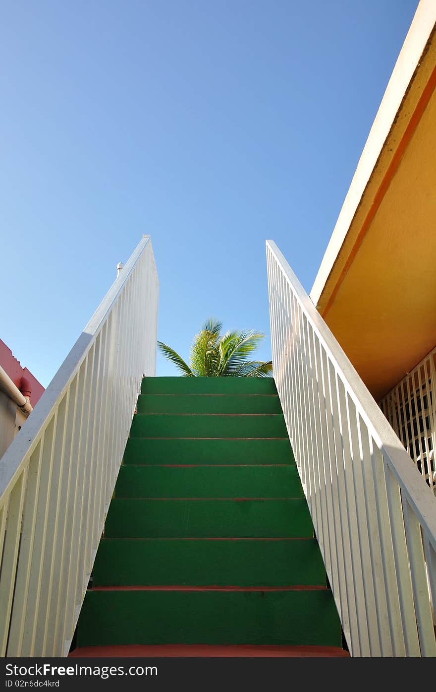 On a clear blue day a side stairway that leads to the roof, typical to most Puerto Rican/Caribbean houses seems as if it is leading to the skies. The top of a palm tree is at first glance. The colors of the house are very tropical; green, pink, orange and white. On a clear blue day a side stairway that leads to the roof, typical to most Puerto Rican/Caribbean houses seems as if it is leading to the skies. The top of a palm tree is at first glance. The colors of the house are very tropical; green, pink, orange and white.