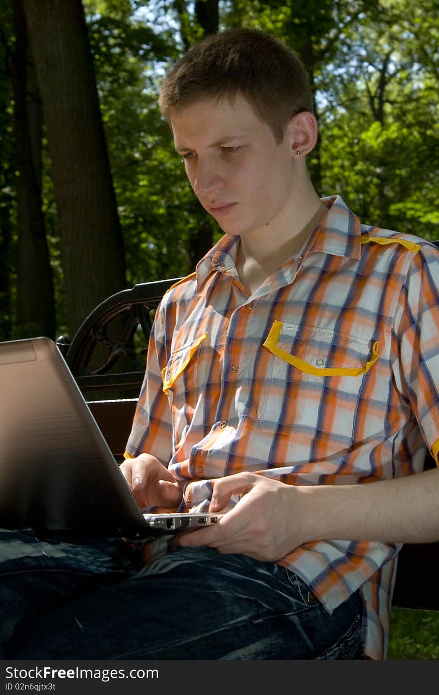 Young man sitting on a bench in green park and types on laptop. Young man sitting on a bench in green park and types on laptop