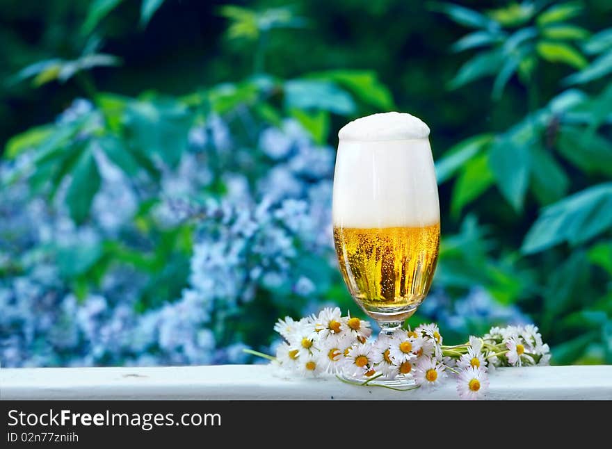 Glass of beer with garland of daisies, standing on the terrace in front of the garden. Glass of beer with garland of daisies, standing on the terrace in front of the garden