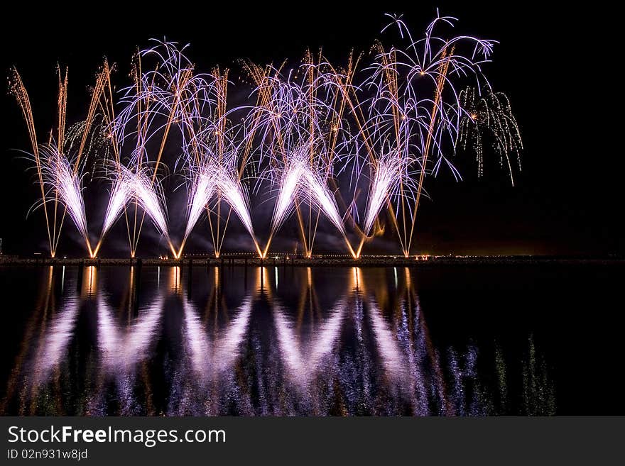 Long exposure photo of multiple fireworks over a lake, against a black sky.